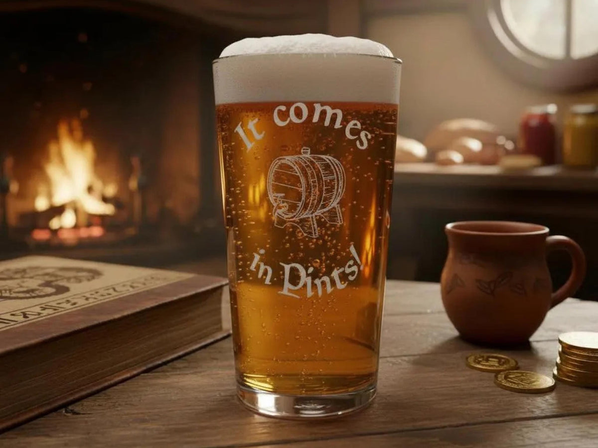 Pint of beer in a glass engraved "It comes in Pints!" with a barrel, on a wooden table by a fire