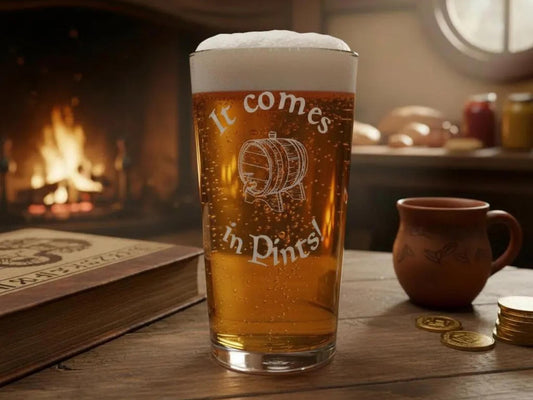 Pint of beer in a glass engraved "It comes in Pints!" with a barrel, on a wooden table by a fire