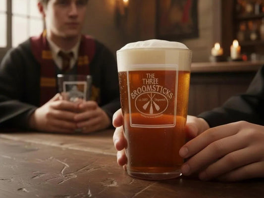 Pint glass of amber beer engraved with The Three Broomsticks pub sign, held on a tavern table