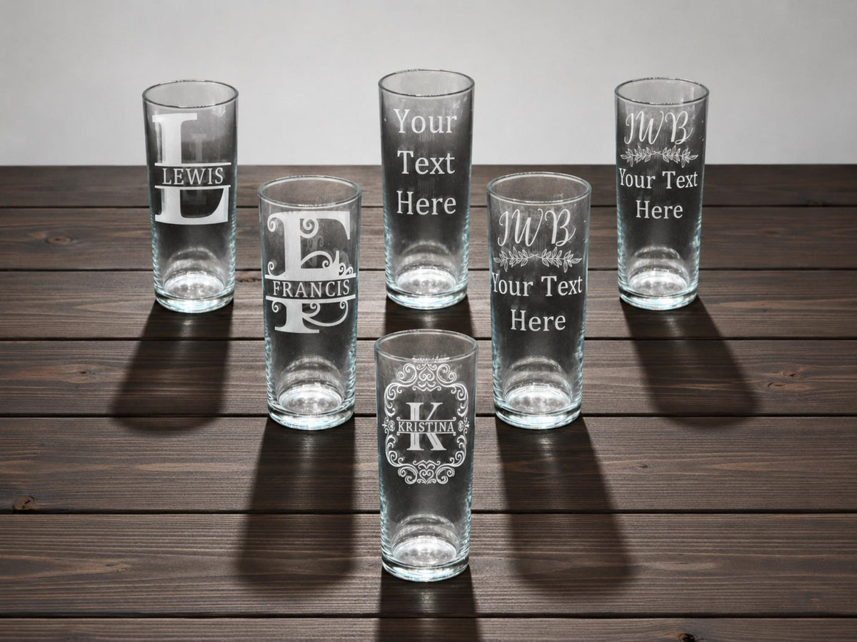 Six tall highball glasses engraved with monogram initials and names like Lewis and Francis, on a dark wooden table