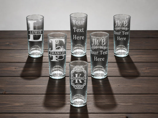 Six tall highball glasses engraved with monogram initials and names like Lewis and Francis, on a dark wooden table