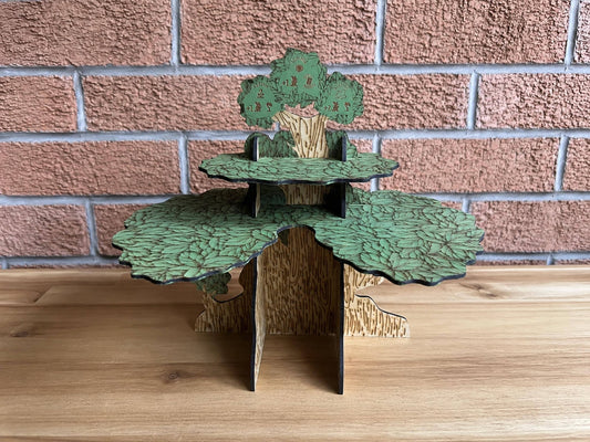 Painted wooden Everdell tree with engraved green leaves and bark, standing on a wooden shelf