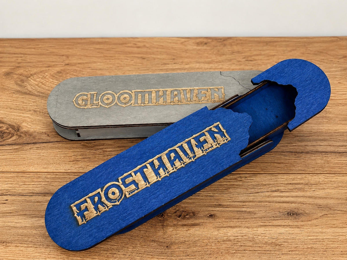 Grey wooden pencil box engraved Gloomhaven beside an open blue pencil box engraved Frosthaven, on a wooden table