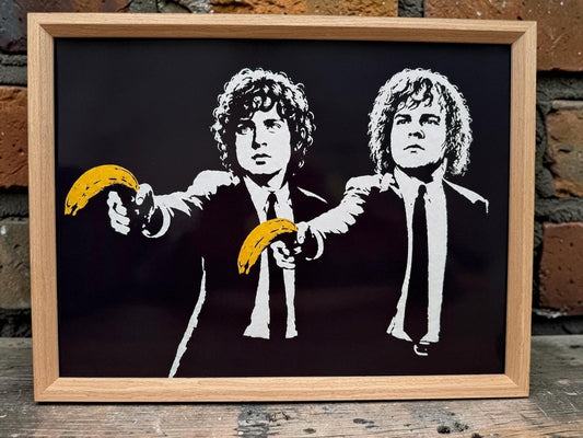 Oak framed black and white street art print of two hobbits in suits holding yellow bananas, against a brick wall